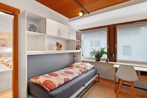 Il s’agit d’une petite pièce comprenant un lit, un bureau et une chaise. On y trouve des étagères au-dessus du lit et une fenêtre avec des rideaux.