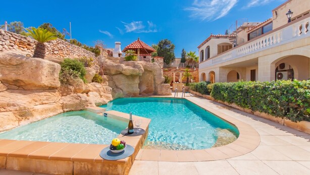 Une piscine avec un ensemble rocheux, un espace de détente avec des boissons, et un bâtiment surmonté d’un toit en tuiles rouges.