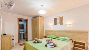 Une chambre à coucher comprenant un lit, des tables de chevet, un bureau, une armoire, un ventilateur, un luminaire et une œuvre d’art accrochée au mur.