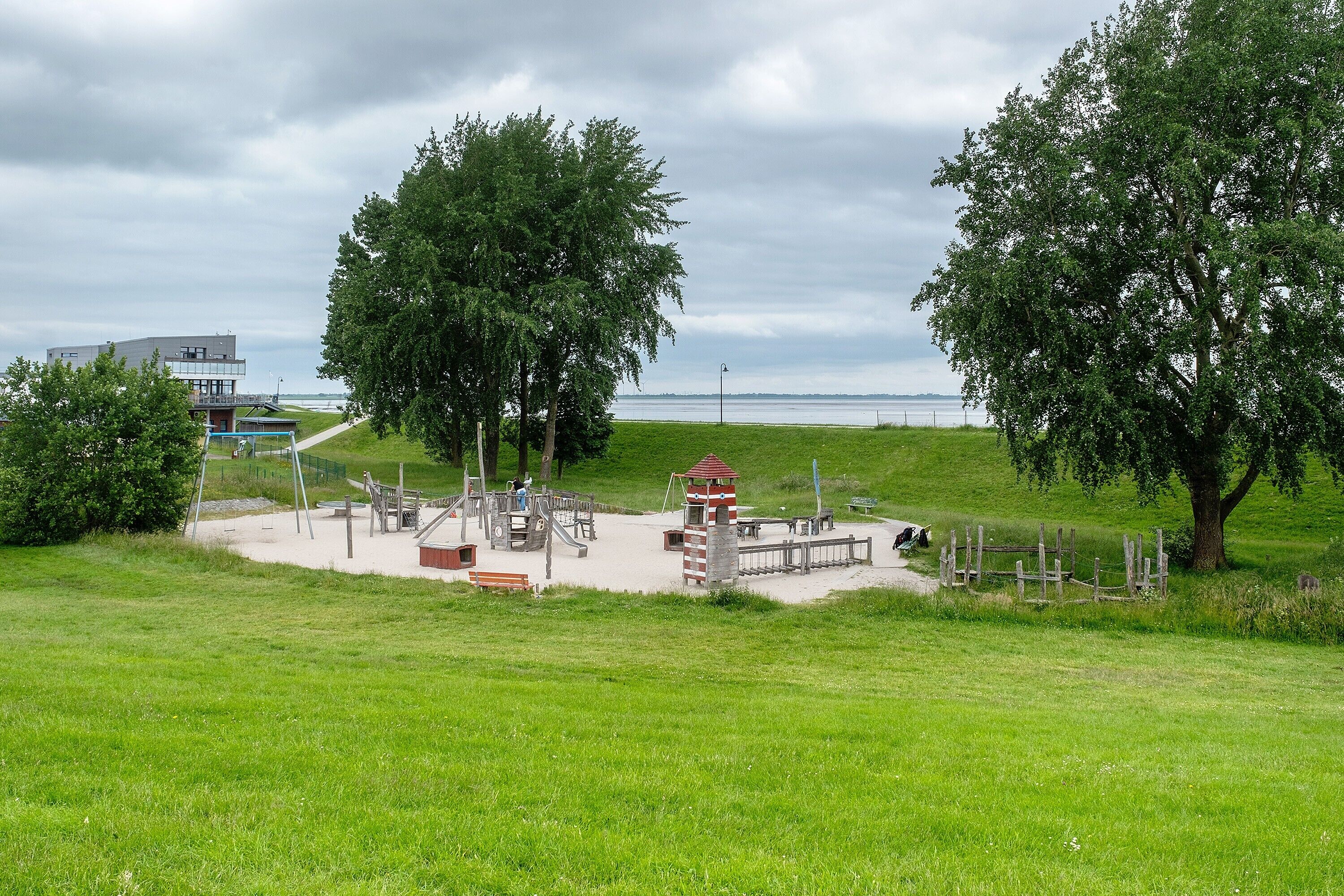 Une aire de jeux avec des balançoires, un toboggan et une structure d’escalade, le tout entouré d’arbres et d’un champ d’herbe.