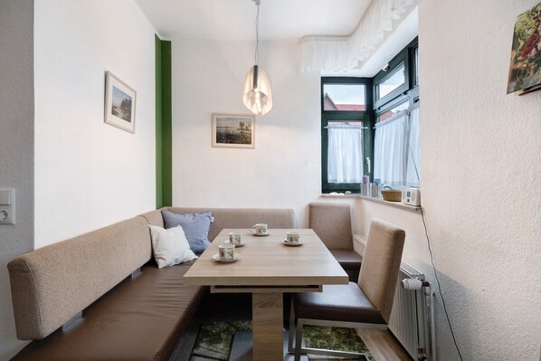 Une pièce comprenant un canapé, une table avec des tasses, un luminaire suspendu, une fenêtre avec des rideaux et un radiateur.