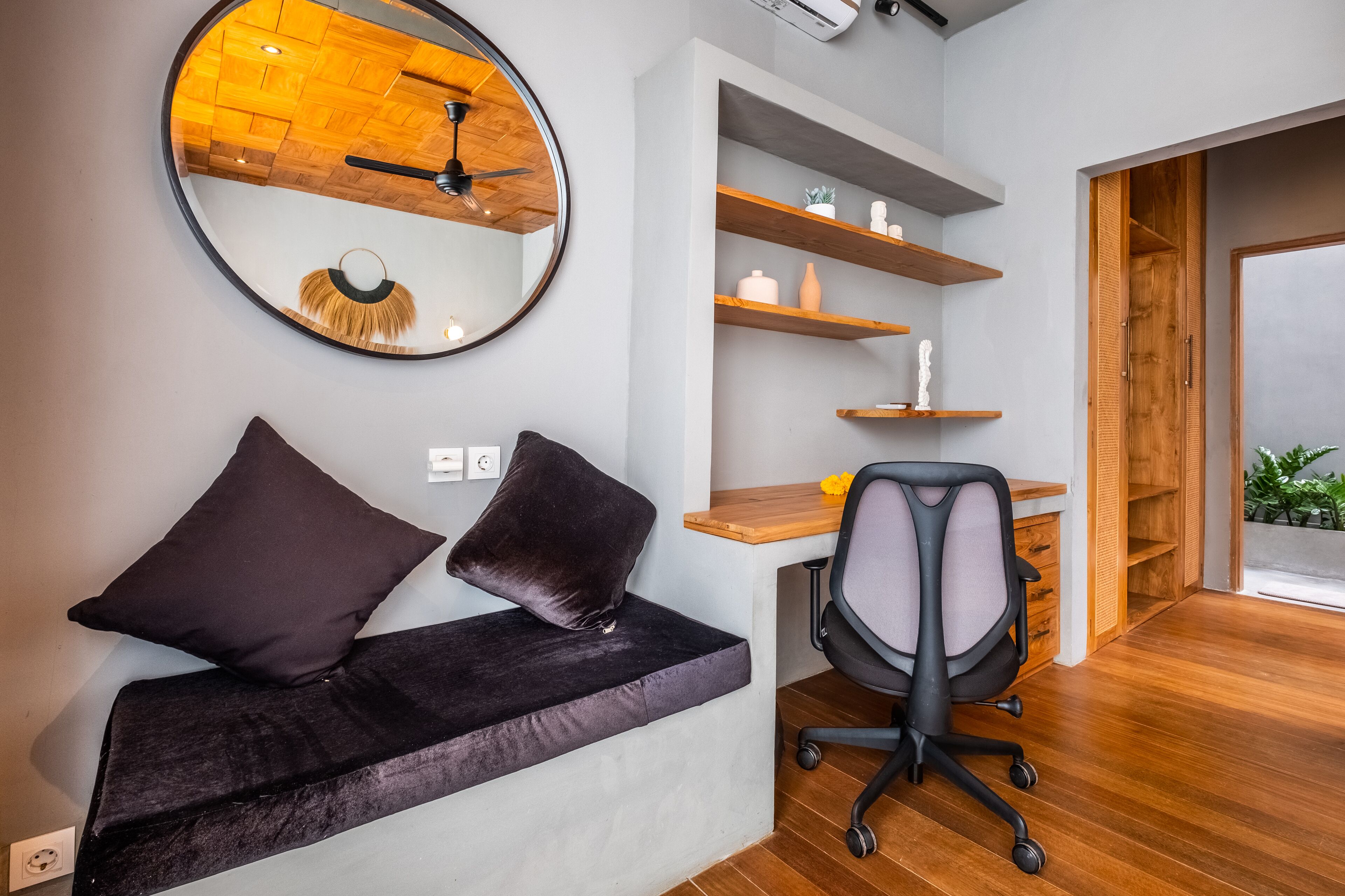 A modern office space with a desk, chair, and a cushioned bench with two pillows. There are wooden shelves above the desk and a large mirror on the wall.