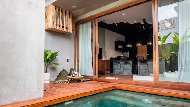 A modern pool area with a wooden deck, a dining area with chairs, and a kitchen visible through glass doors. - Hebe Villa by Betterplace (Canggu)