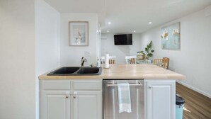 A modern kitchen with a wooden countertop, white cabinetry, a built-in dishwasher, and a TV mounted on the wall. - Coastal Cozy and Close to Beach with Turf Yard! (Carolina Beach)