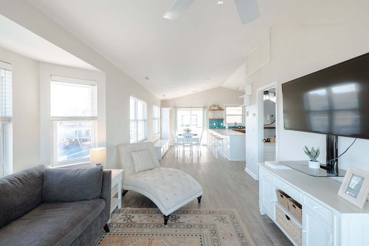 Amplia sala de estar con un sofá gris, un sillón blanco, un televisor sobre un mueble blanco y una cocina al fondo.