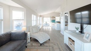 A spacious living area with a grey sofa, a white armchair, a TV on a white cabinet, and a kitchen in the background. - Ocean Views + Across from Beach Access! (Surf City)