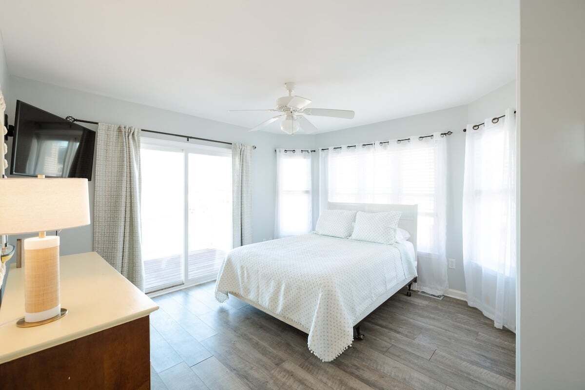 A bedroom with a bed, a ceiling fan, a TV on the wall, and a window with curtains.