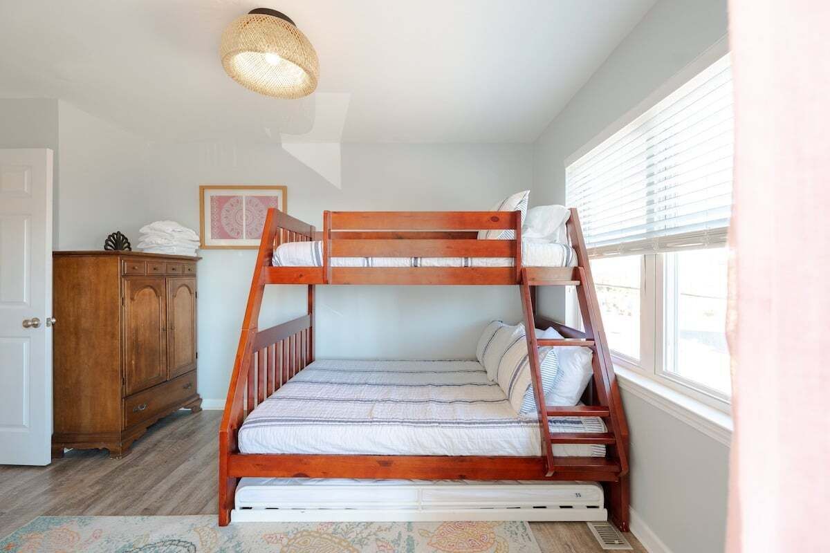 Un letto a castello con struttura in legno, un materasso bianco e cuscini. A sinistra, una cassettiera di legno e, a destra, una finestra con persiane.