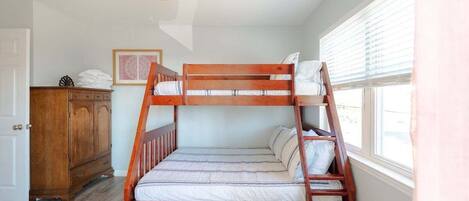 Un letto a castello con struttura in legno, un materasso bianco e cuscini. A sinistra, una cassettiera di legno e, a destra, una finestra con persiane.