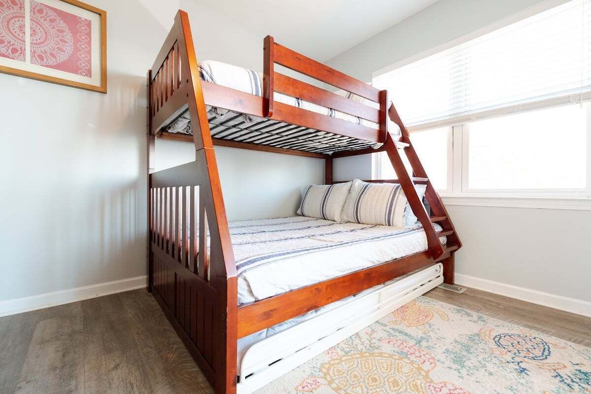 A bunk bed with a wooden frame and white bedding, a patterned rug, and a window with blinds.