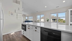 A modern kitchen with white cabinetry, a built-in oven, and a dishwasher. There is a window providing a view of the outside. - Ocean Views + Across from Beach Access! (Surf City)