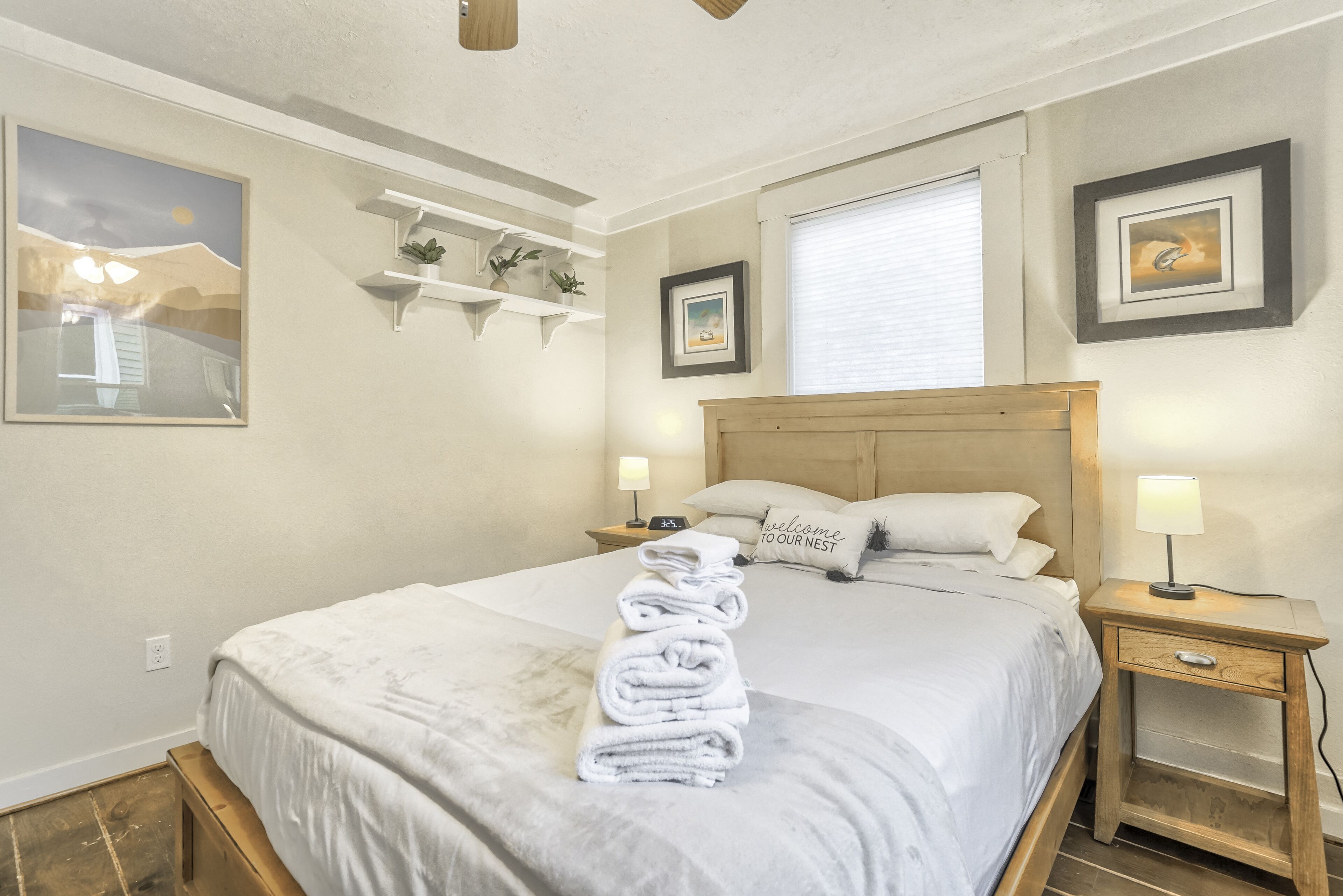 A neatly made bed with a folded towel, a wooden headboard, bedside lamps, and wall-mounted shelves with plants.