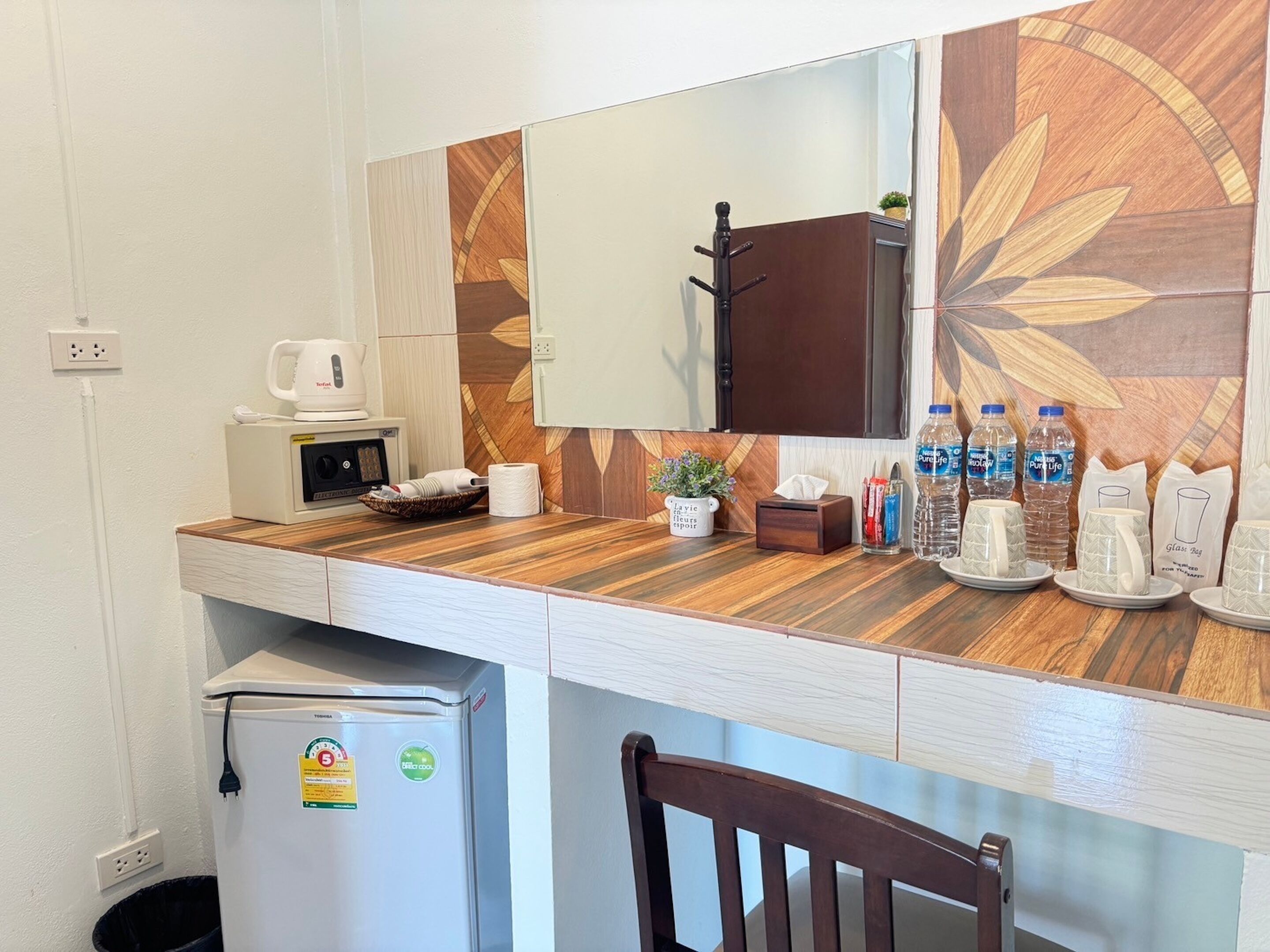 Eine Küchenzeile mit Mikrowelle, Wasserkocher und kleinem Kühlschrank. Ein Holztisch mit Spiegel darüber, und ein Stuhl ist im Vordergrund zu sehen.