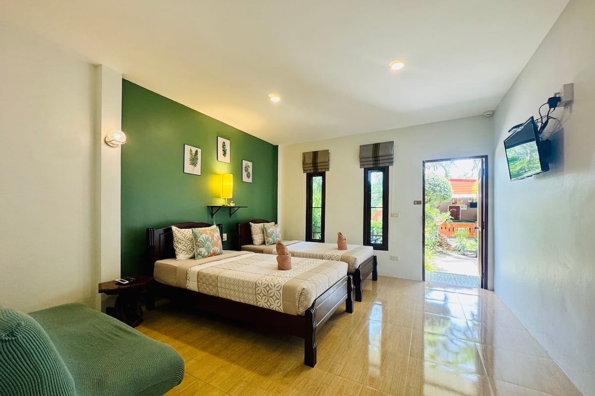 Habitación de hotel con dos camas, una pared verde, una pequeña mesa, televisión y vistas al exterior.