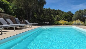 Uma piscina retangular com água azul cristalina, rodeada por um deck de madeira e cadeiras de descanso.
