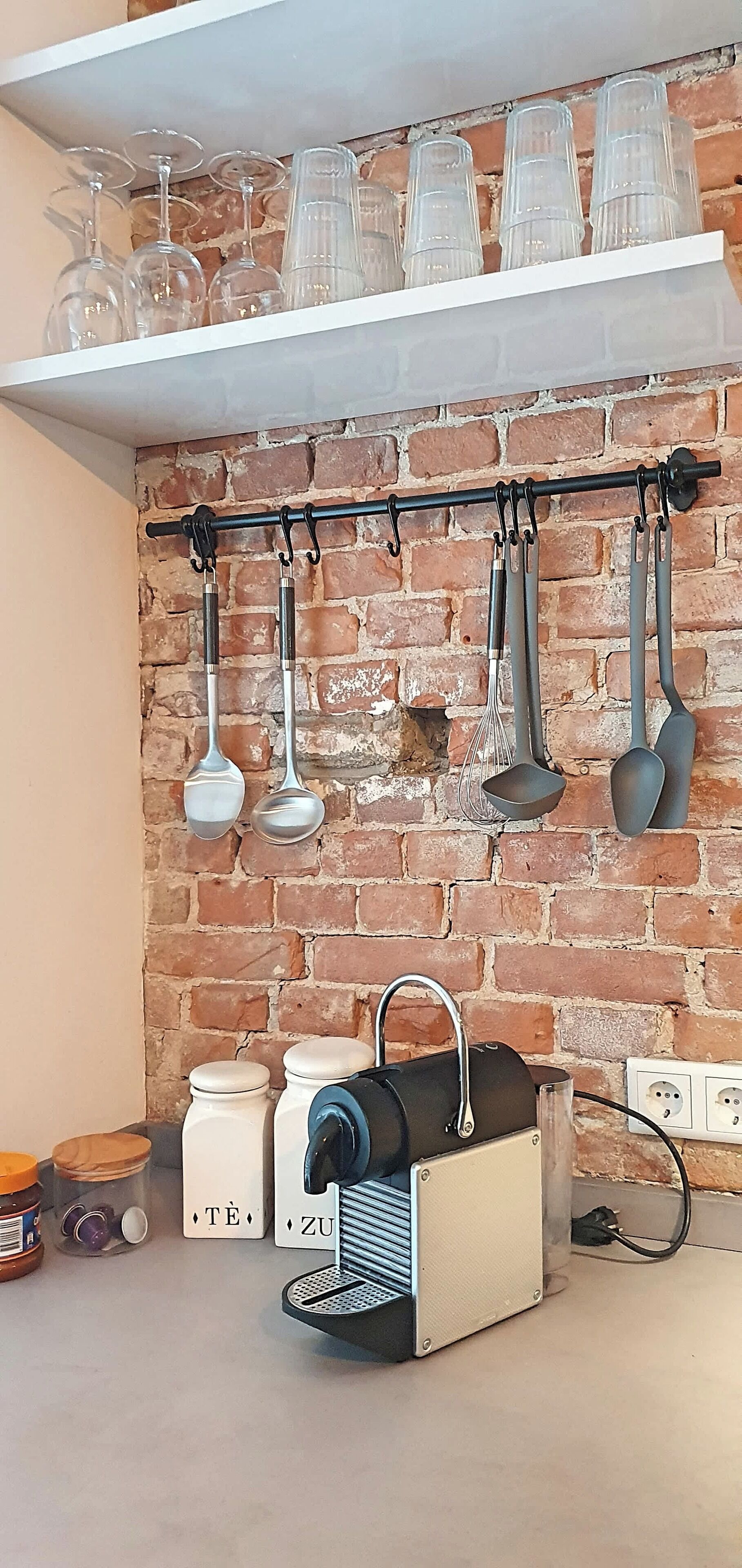 Una cocina con pared de ladrillo, una cafetera, frascos etiquetados 'TÊ' y 'ZU', y utensilios colgando.