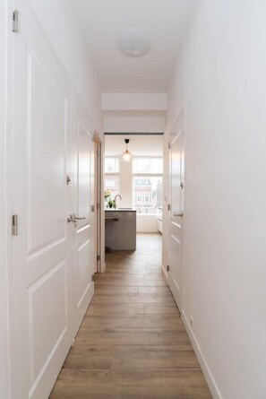 Un couloir étroit, avec des portes blanches et un plancher en bois, mène à une cuisine dotée d’une fenêtre et d’un luminaire suspendu.
