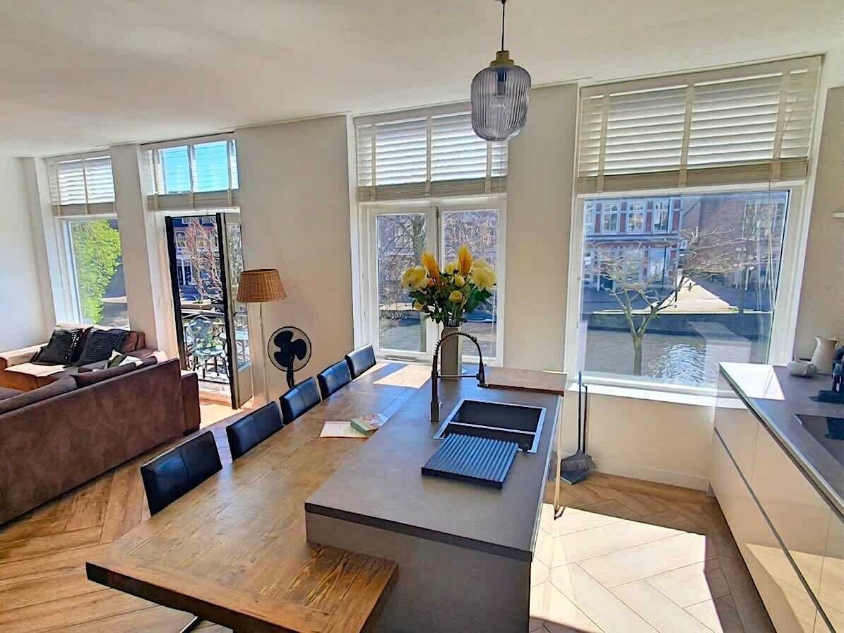 Una sala moderna con una mesa de comedor de madera, un sofá, un ventilador y vistas a un río a través de amplios ventanales.