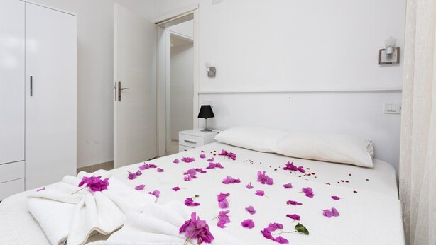 A hotel room with a bed covered in white linens and purple flowers, a white wardrobe, a bedside table with a lamp, and a door leading to another room. - Villa Casa Mia in Dalyan (Ortaca)