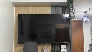 A modern living room with a flat-screen TV mounted on a wooden wall, a small white shelf with decorative items, a black floor lamp, and a cabinet with a glass door. - A cozy home in the middle of the city (Cebu City)