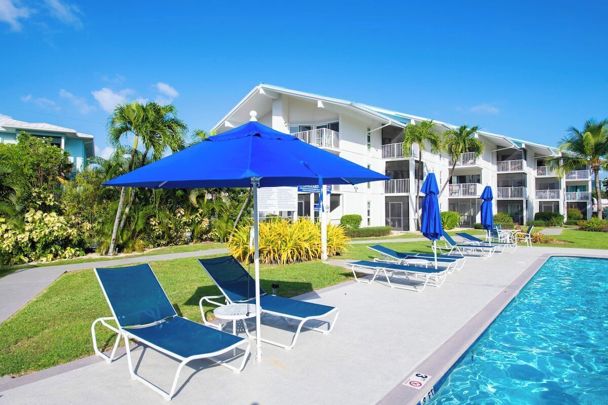 A pool area with lounge chairs and umbrellas, a clear sky, and a building with balconies.