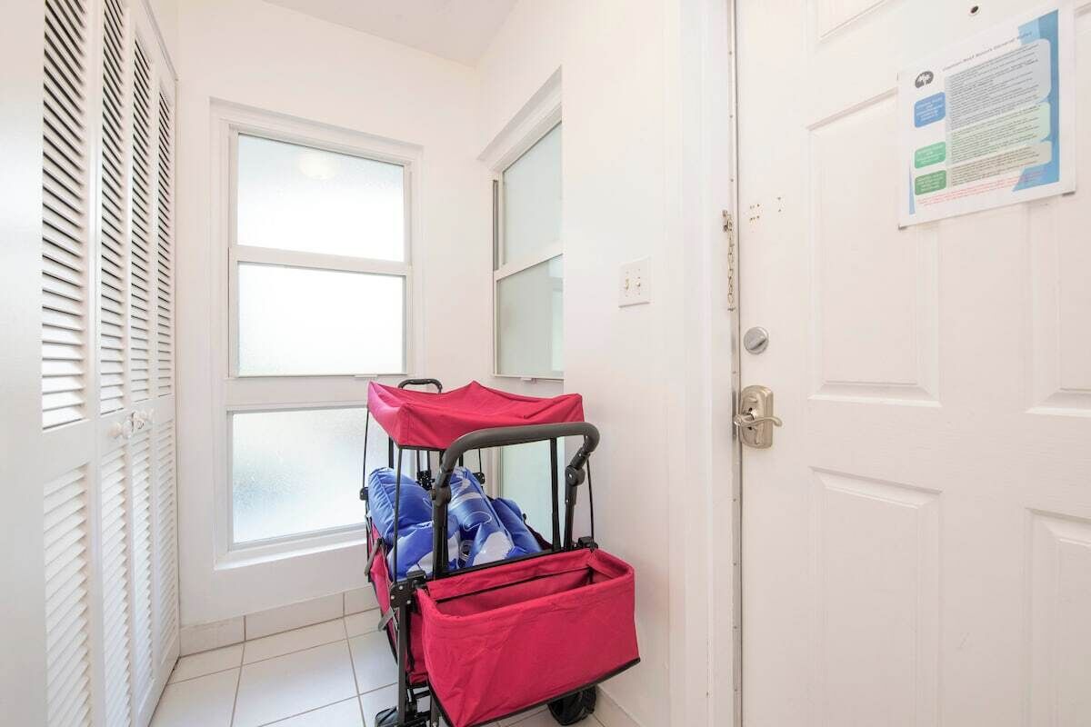 A hallway with a white door, a window with blinds, and a red luggage cart.