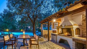 An outdoor patio with stone flooring, a wooden table with chairs, a barbecue grill, and a pool in the background. - Villa Alkyon in Paxi (Paxos)