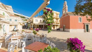 An outdoor café area with tables and chairs, surrounded by buildings with balconies and a church with a bell tower. - Villa Alkyon in Paxi (Paxos)