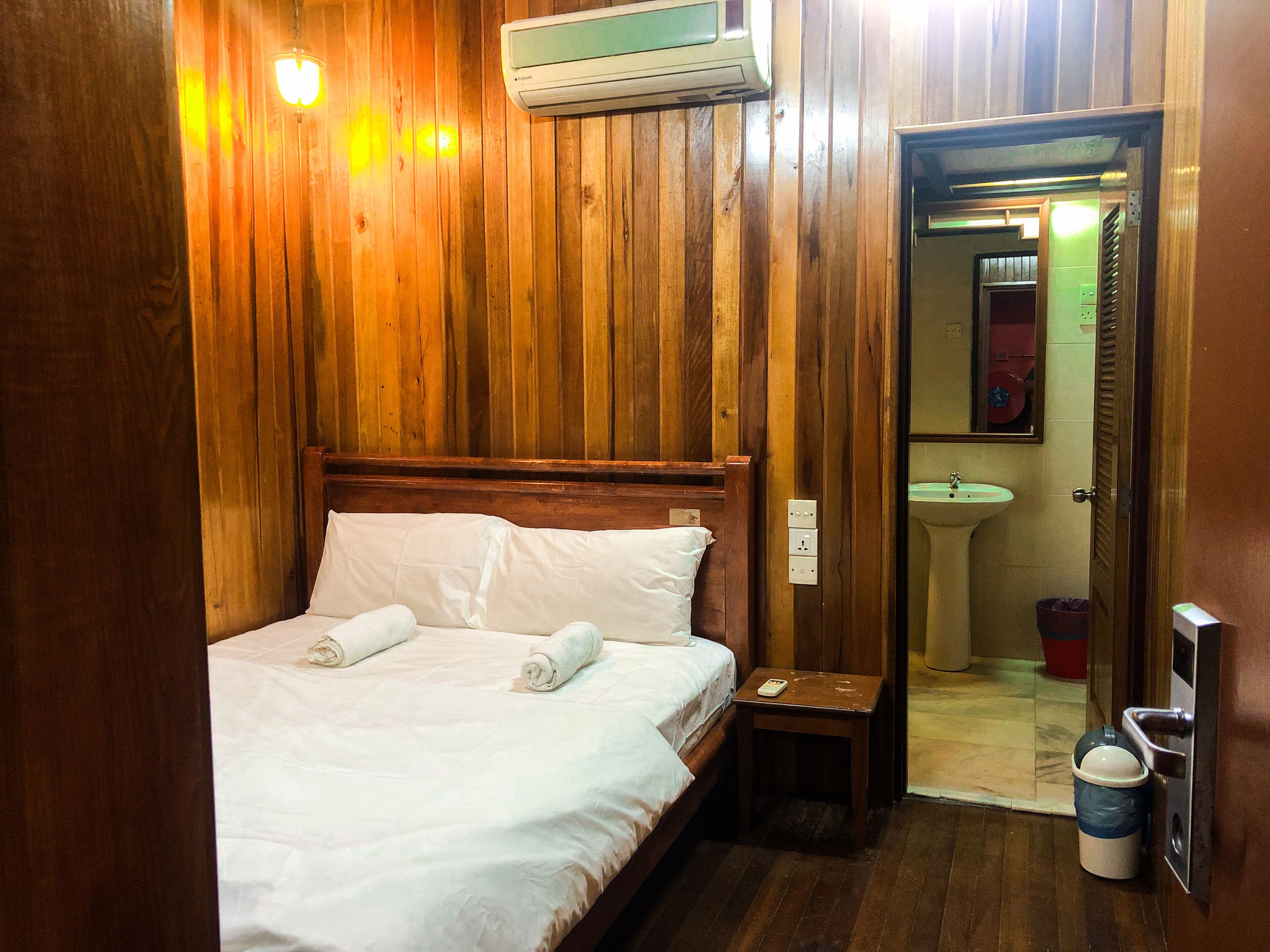 Une pièce aux murs lambrissés, comprenant un lit, une table de chevet, un climatiseur fixé au mur, une salle de bain avec un lavabo et un miroir, et une poubelle.