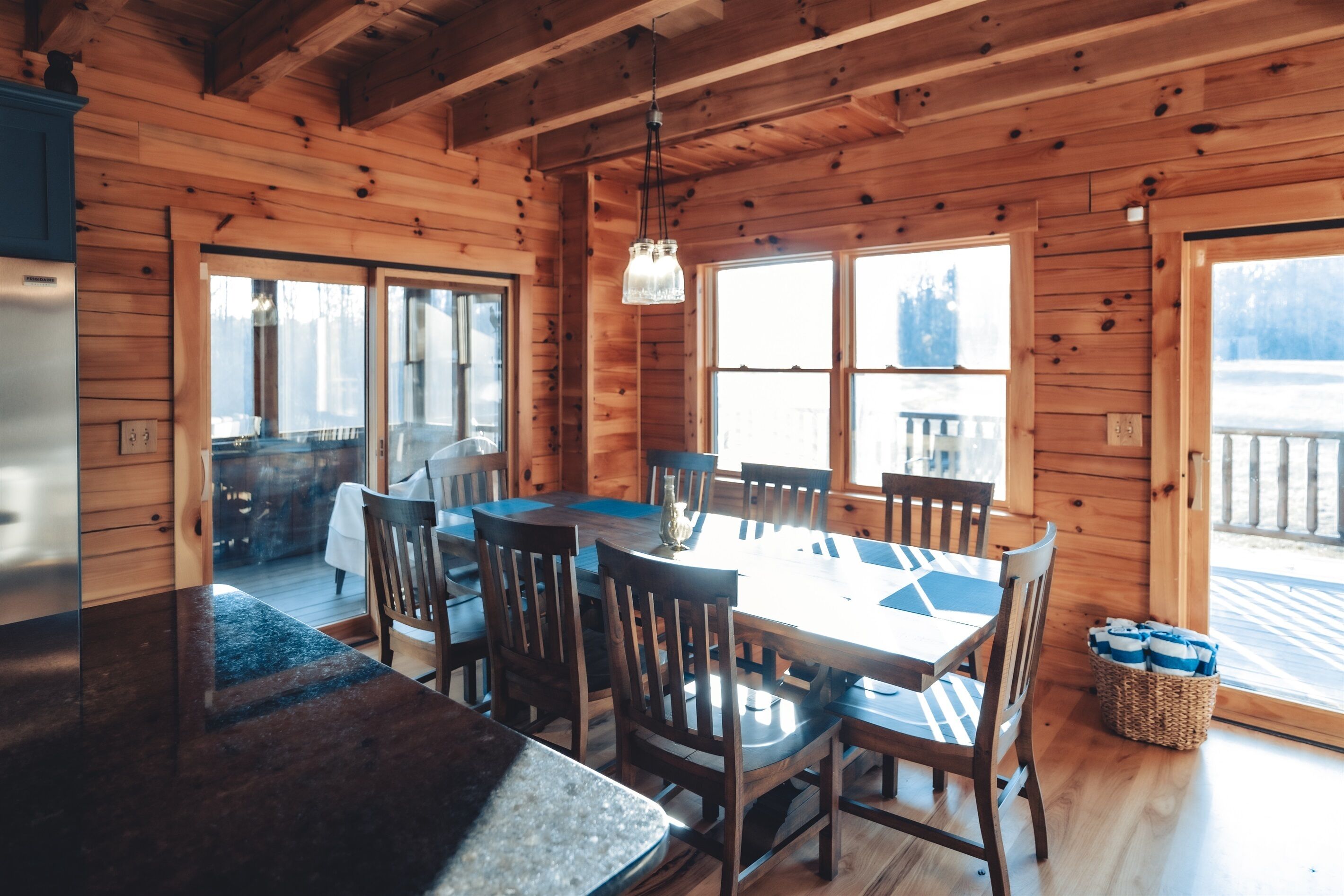 A wooden cabin interior with a dining table, chairs, a refrigerator, and large windows.