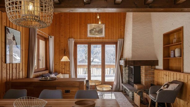 A cozy wooden interior with a dining area, fireplace, and large windows overlooking a snowy landscape.