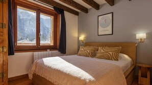 A bedroom with a wooden bed, a window with curtains, a wall-mounted lamp, and a framed picture.