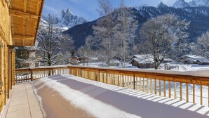 A wooden balcony with a snow-covered ground, overlooking a mountainous landscape with snow-covered houses.