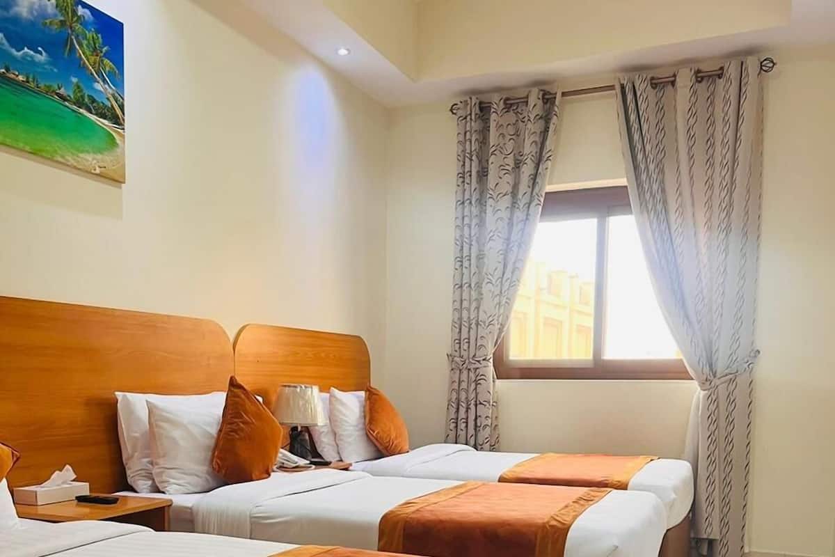 Une chambre d’hôtel avec deux lits, chacun doté d’une tête de lit en bois, de draps blancs et de coussins décoratifs orange. On y trouve une fenêtre avec des rideaux à motifs, un tableau représentant une scène de plage accroché au mur et un sol carrelé.