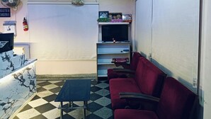 Zona de recepción con mostrador de check-in, área de espera con sillas rojas, un televisor en un estante y un reloj en la pared.