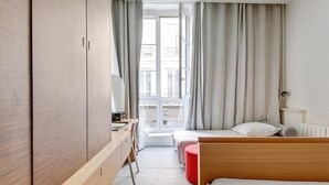 A modern hotel room with a wooden wardrobe, a desk with a lamp, a bed with a red ottoman, and a view of the city through large windows.