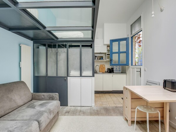 Un espacio habitable compacto con sofá, un escritorio pequeño de madera, kitchenette y puerta corrediza de vidrio que da a un balcón.