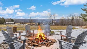 A fire pit with four Adirondack chairs arranged around it, set in an outdoor area with a view of distant hills and a clear sky.