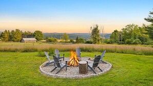 A circular fire pit with four Adirondack chairs around it, set in a grassy area with a view of distant hills and a barn.