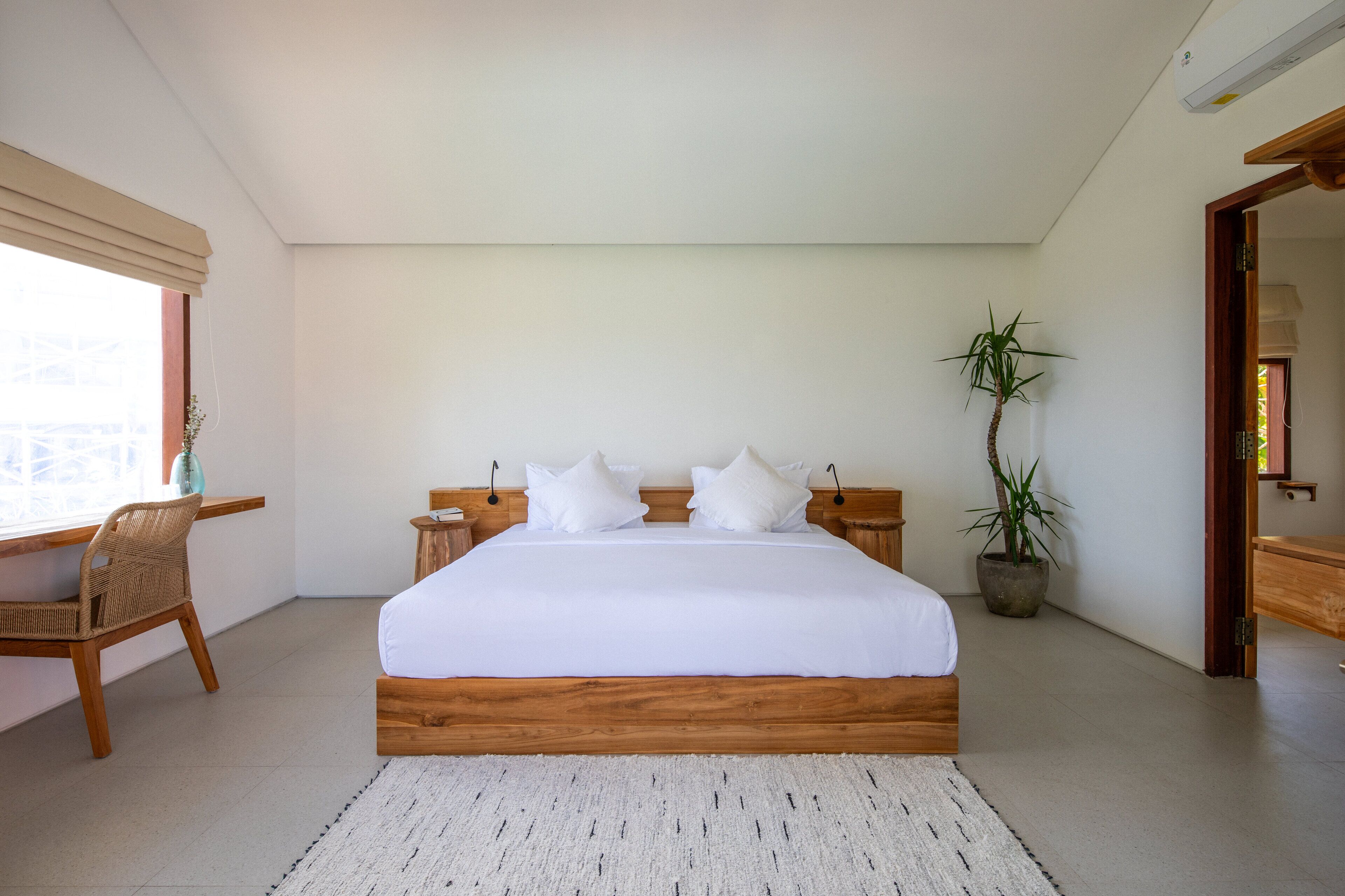 A bedroom with a large bed, wooden headboard, white bedding, a small table with a chair, a potted plant, and a window with blinds.