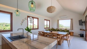 Une cuisine moderne avec un plan de travail en marbre, une table à manger en bois et des chaises. De grandes fenêtres offrent une vue sur la verdure environnante.