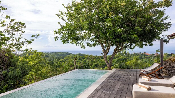 Une piscine à débordement avec une terrasse en bois, des chaises longues et une vue sur l’océan et la forêt.