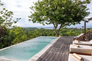 An infinity pool with wooden deck, lounge chairs, and a view of the ocean and forest.