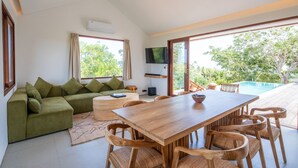 Un salon moderne avec une table à manger en bois, un canapé vert et de grandes fenêtres donnant sur l’extérieur.
