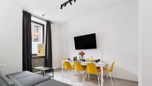 Una sala moderna con un sofá gris, una mesa de comedor con sillas amarillas, un televisor de pantalla plana montado en la pared y una ventana con cortinas negras.
