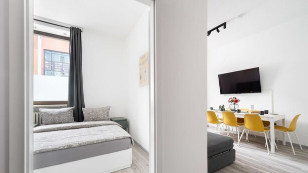 Un dormitorio moderno con cama, televisor y zona de comedor con sillas amarillas.