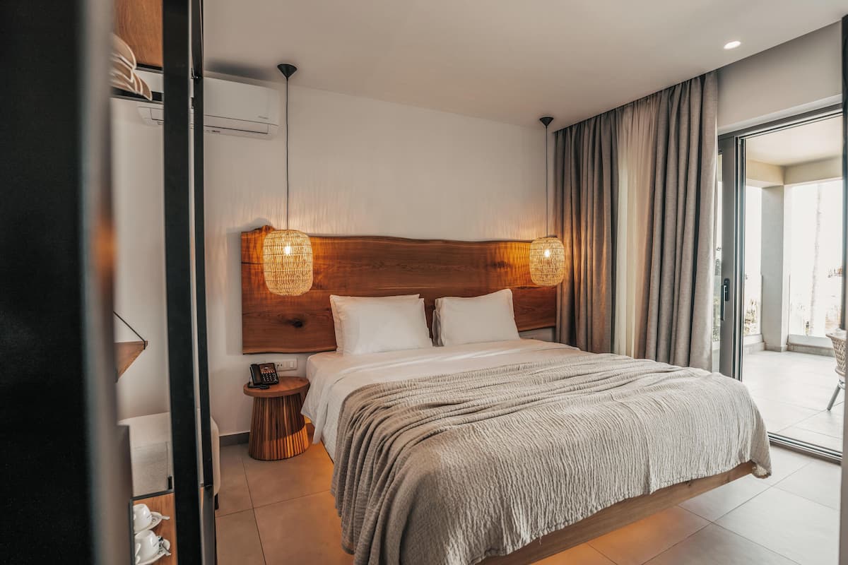 A modern hotel room with a large bed, two bedside lamps, a small side table, and a view of the outside through a glass door.