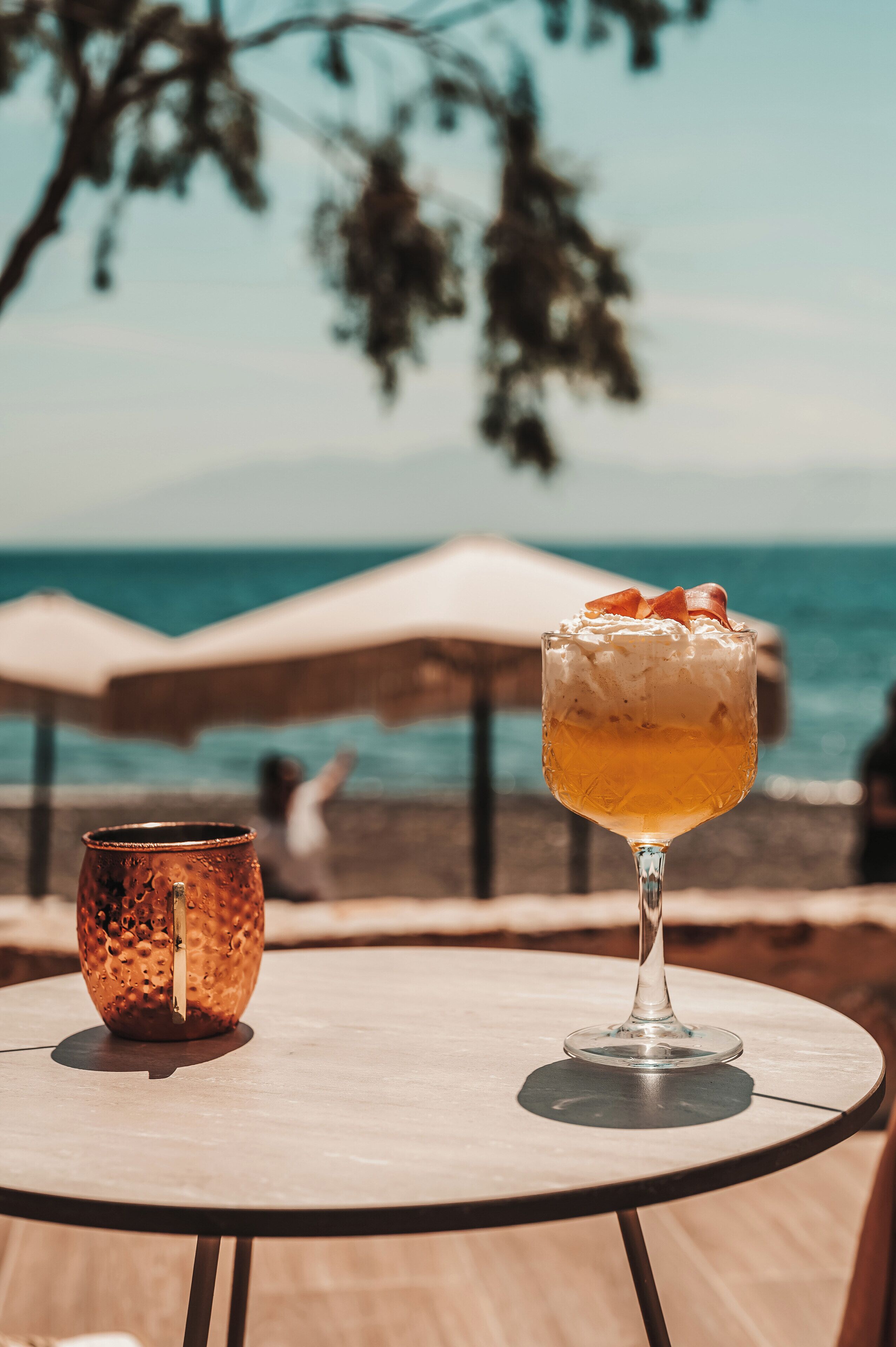 A table with a glass of cocktail and a copper mug, set against a beach backdrop with umbrellas and the sea.