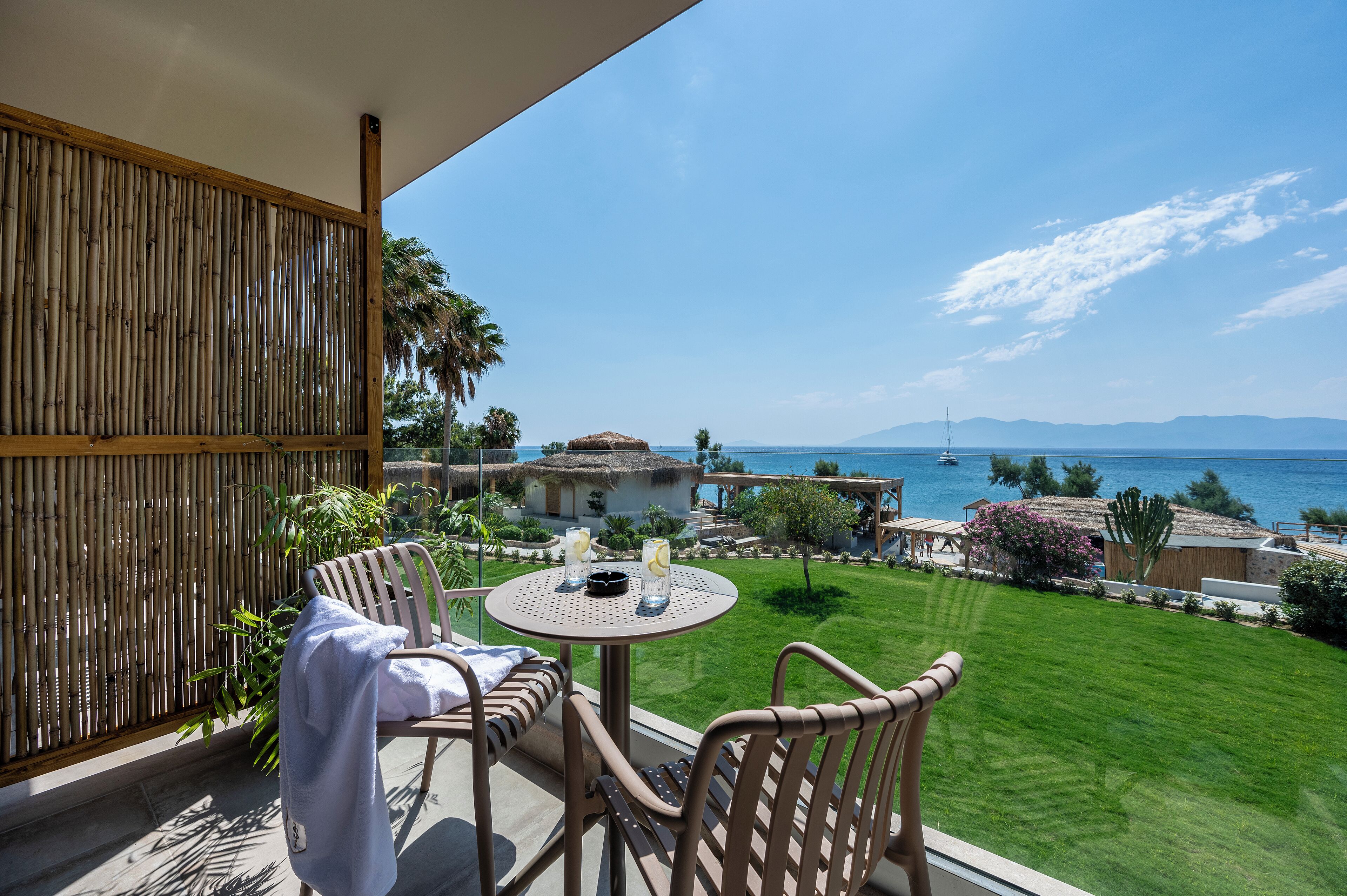 Een balkon met rieten meubilair, een ronde tafel met drankjes, en uitzicht op zee en bergen.