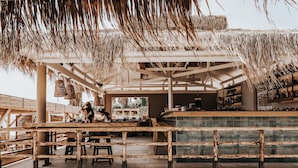A beachside bar with wooden tables and chairs, a thatched roof, and a view of the ocean. - Sagredo Beachfront Retreat & Spa by E-GEO (Kos)